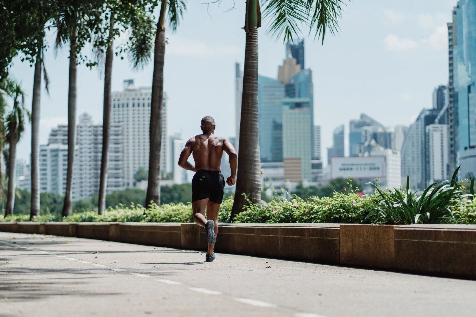A shirtless man jogging on a sunny day in an urban cityscape with tall buildings.